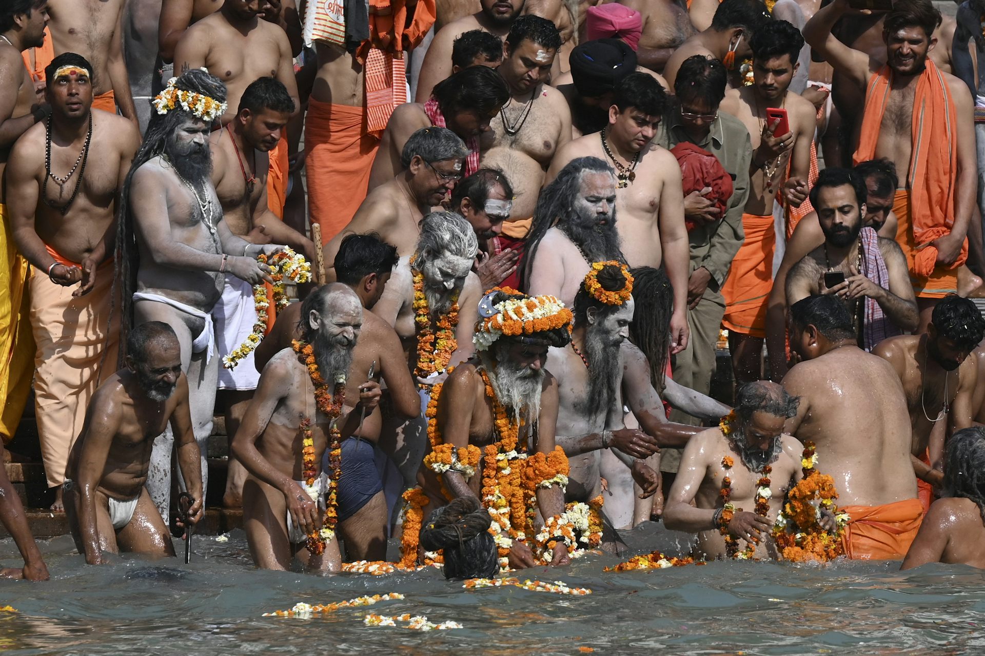 Men are shoulder to shoulder on the edge of the water. Most are naked from the waist up. Many wear flowers and orange clothing.